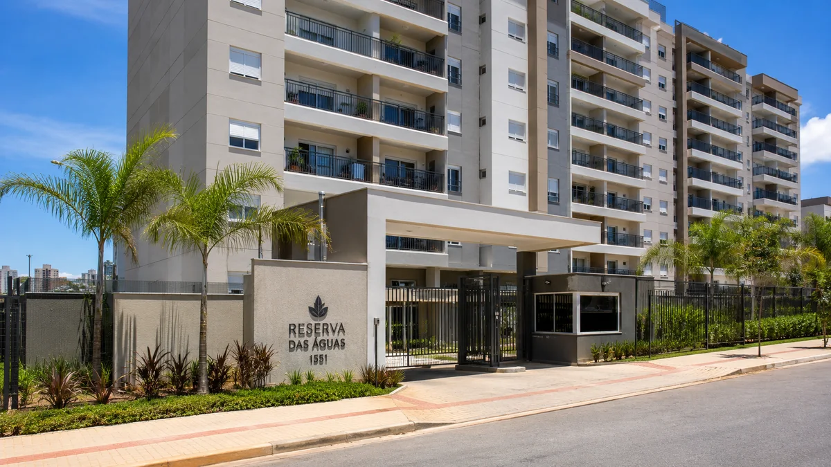 Apartamentos e condomínios. Fachada de um condomínio de apartamentos residenciais moderno com sacadas de vidro e portaria, cercado por árvores em um dia ensolarado no Brasil